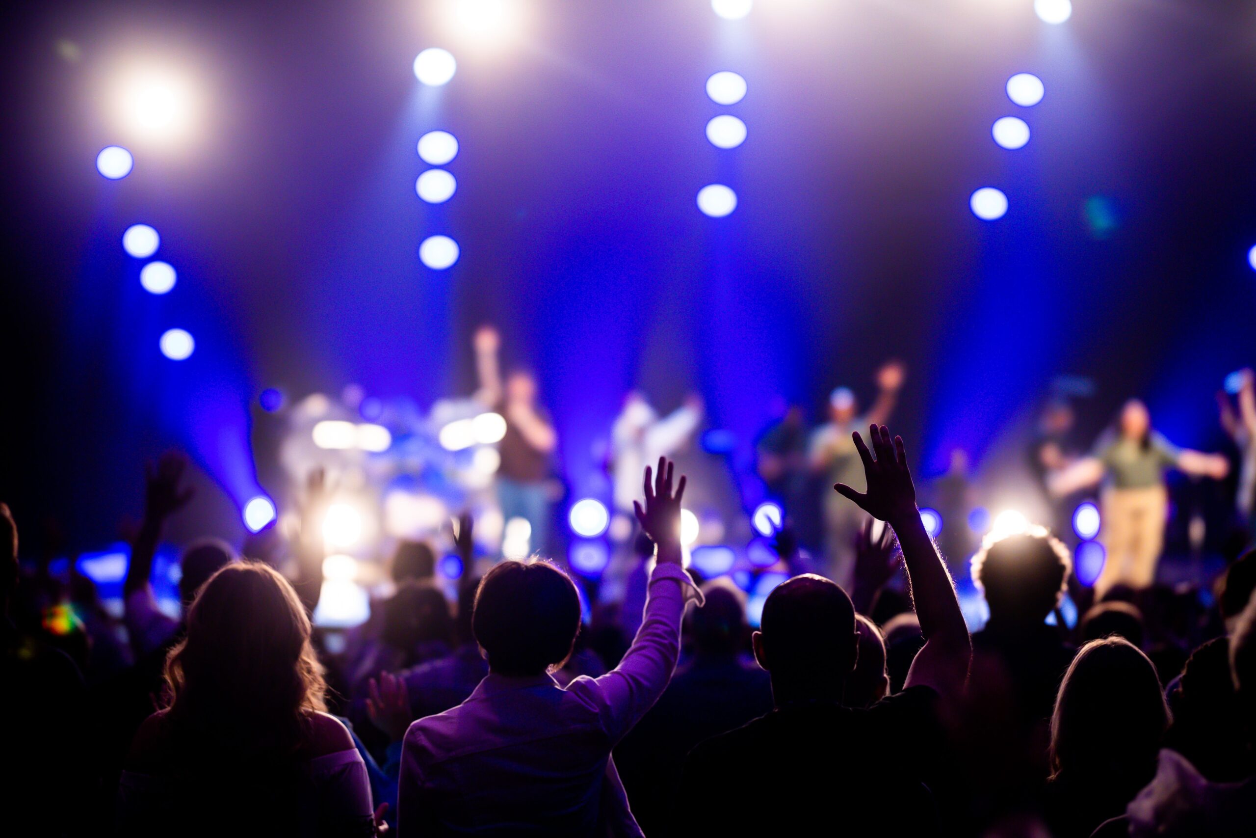 Crowd at a concert with raised hands and blue stage lights in the background.